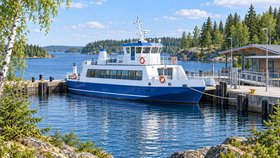 Sinivalkoinen matkustaja-alus on kiinnitetty köysillä laituriin. Aluksen vieressä on matala puurakennus ja laiturikaiteita. Satamaa ympäröivät sininen vesi, kalliorannat ja havumetsä.