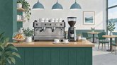 Kahvilan puutiskillä on espressokone, kahvimylly ja kaksi kahvikuppia aluslautasineen. Lasikuvun alla on croissanteja, ja taustalla näkyy pyöreitä pöytiä, vihreitä tuoleja, riippuvalaisimia ja viherkasveja.